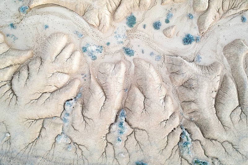 An aerial photograph captures striking erosion patterns in a barren landscape, displaying intricate textures and contrasting hues of sand and sparse vegetation, resembling abstract art