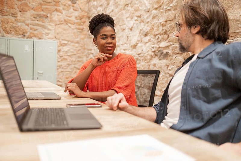 Two professionals in a coworking space engage in discussion, surrounded by laptops and exposed brick decor, highlighting modern and collaborative work environments