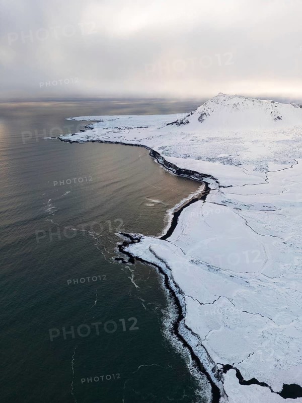Aerial view capturing the stark beauty of Iceland's winter coastline. The snow-covered ground contrasts with the dark waters, showcasing the remote and wild landscape