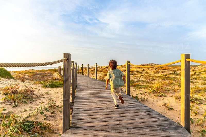 Little boy runs along a wooden boardwalk through sand dunes toward the atlantic beach at golden hour, carefree summer holiday energy and exploration by the sea