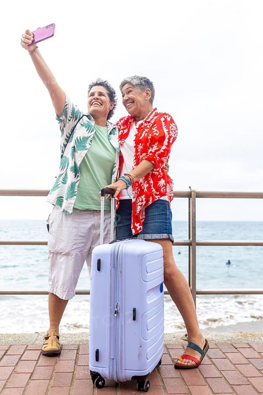 Two mature lesbian women take a selfie together by the seaside, celebrating their summer holiday preparations with a suitcase