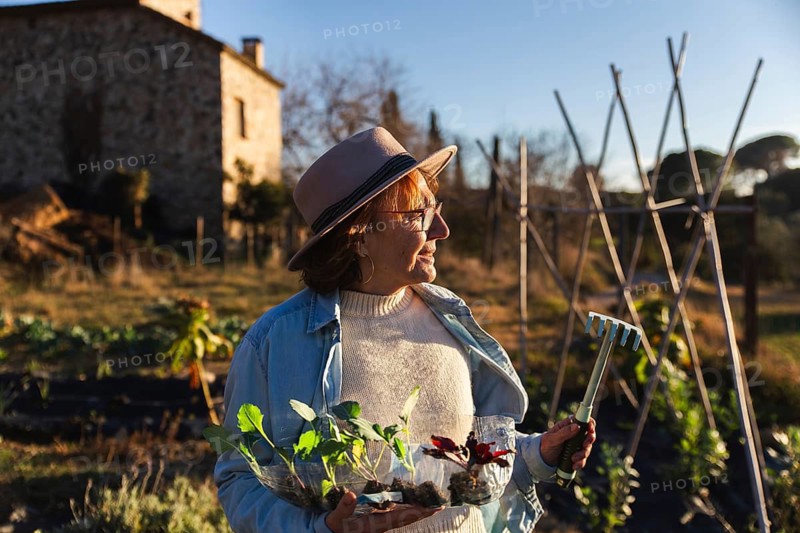 An woman in casual attire and a hat enjoys gardening under the sun, holding seedlings and a mini rake, surrounded by an expansive rural landscape and an old stone building in Barcelona, Spain