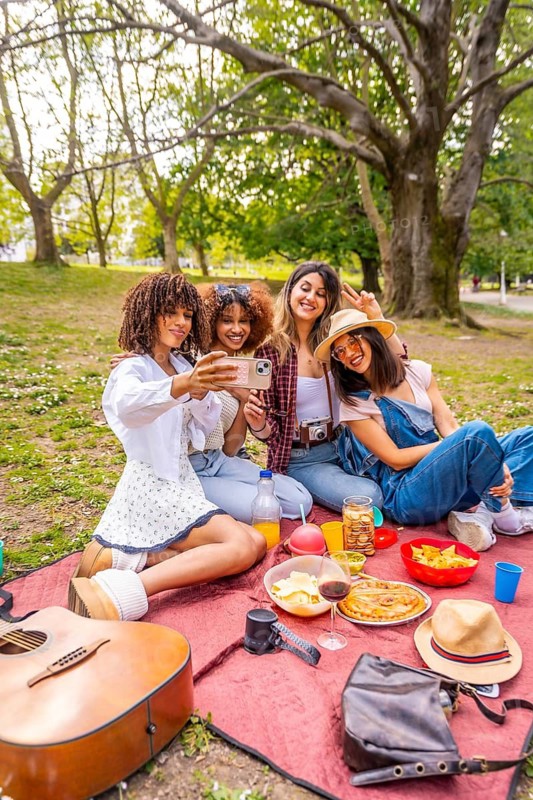 Four female friends taking a selfie with smartphone during a picnic in a park, enjoying music, food and drinks