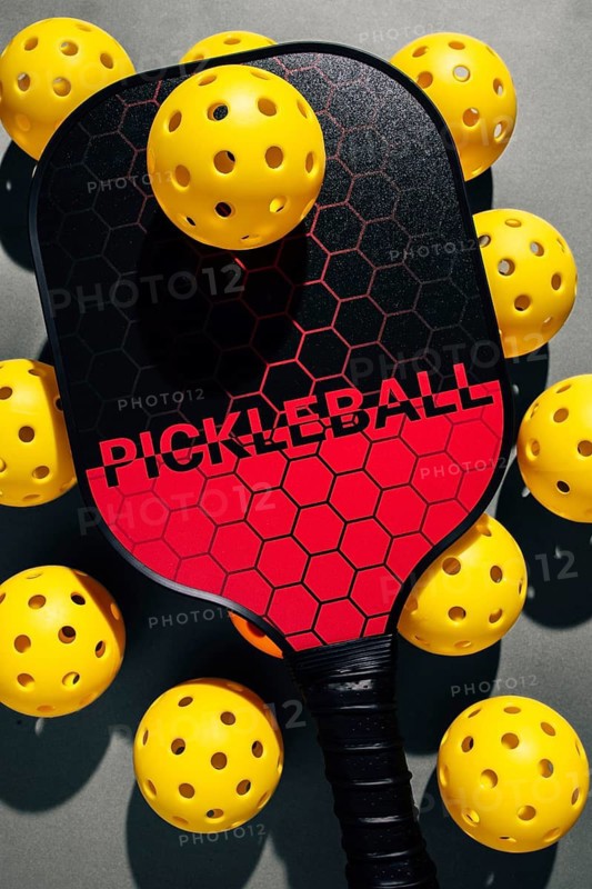 Top view of a vibrant pickleball setup featuring a black and red paddle surrounded by scattered yellow balls on a gray studio background, highlighting modern recreation