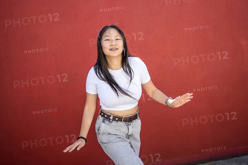 Cute urban female asian artist dancing K-pop dance smiling at camera outdoors against a red wall