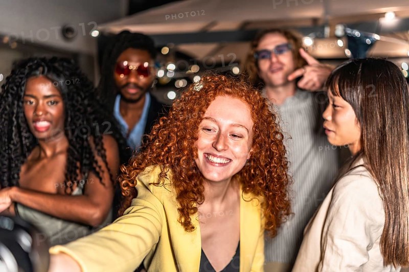 Happy elegant diverse friends taking selfie in a terrace during night party with lights decorating the background