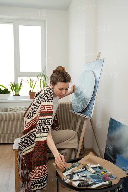 An artist in a cozy, light filled studio works on a canvas with paintbrushes and tubes nearby, creating a serene and focused atmosphere