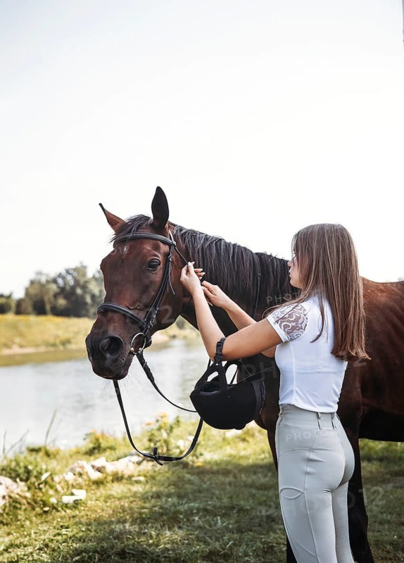 A young girl in equestrian attire prepares her horse by a peaceful river. The intimate connection between human and animal is highlighted in this serene outdoor setting