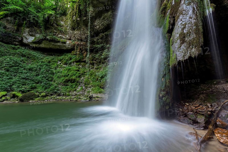 A waterfall flows into a small pool of water. The water is clear and the rocks are scattered around the pool. Lons le Saunier, Jura, Burgundy Franche Comte, France