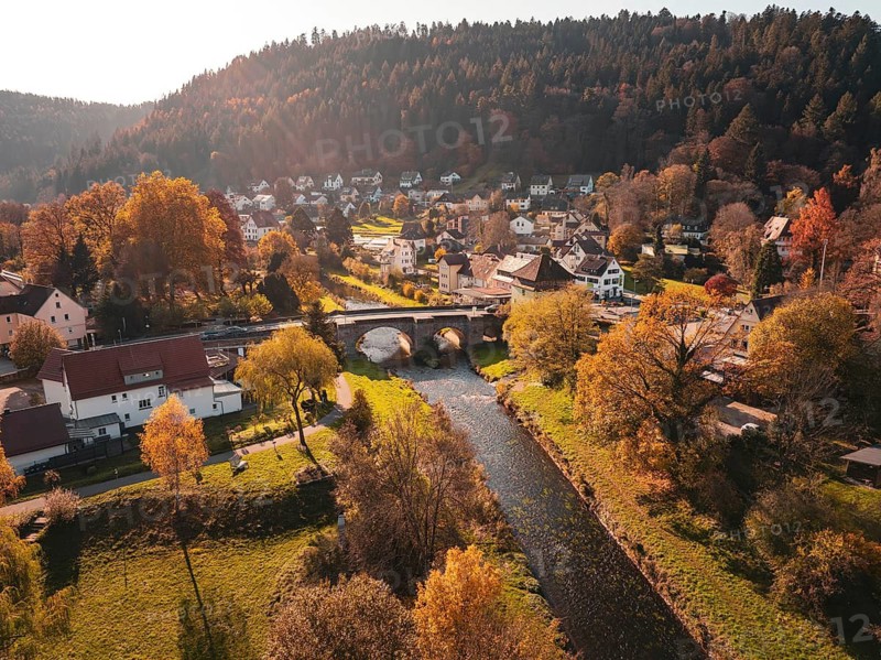 Picturesque autumnal village with river and bridge, surrounded by colourful trees and hills, Calw, Black Forest, Germany