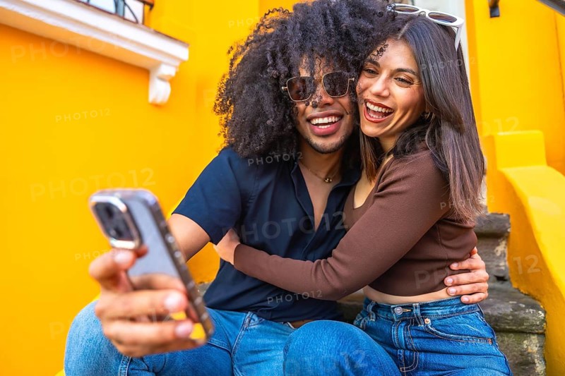 Young couple taking a selfie with a smartphone, hugging and smiling, sitting on colorful steps, enjoying a special moment together