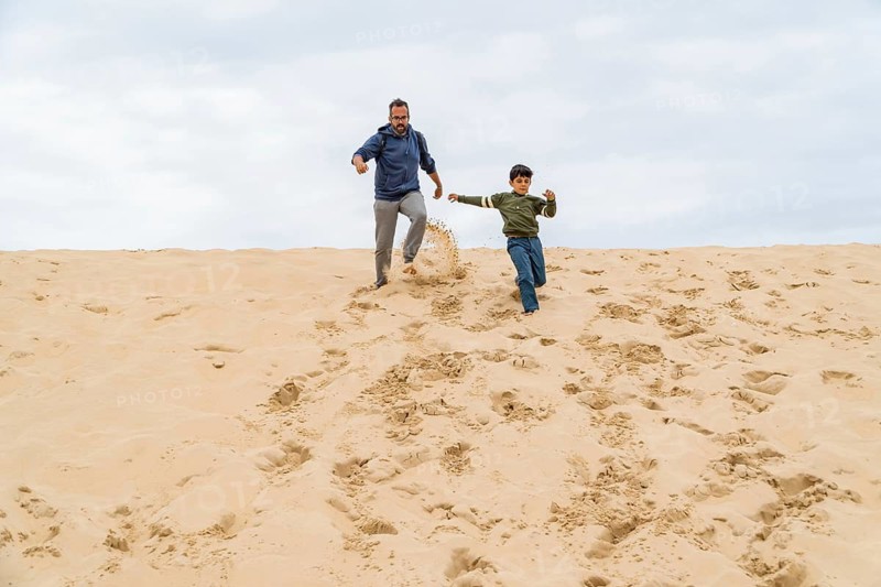 A joyful father and son run down the dune of Pilat, leaving footprints behind. Their laughter fills the air, capturing a moment of freedom and fun on a cloudy day