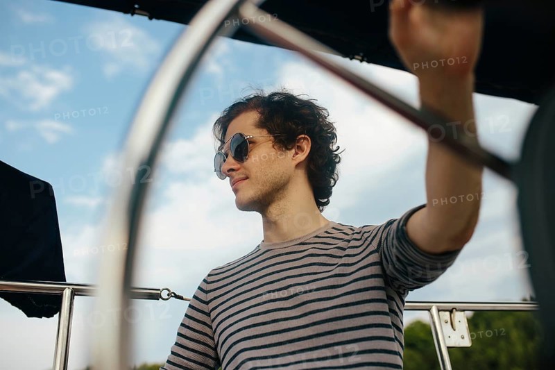 A man enjoys a peaceful afternoon sailing, steering a sailboat under clear skies. The striped shirt and sunglasses reflect a casual, nautical theme