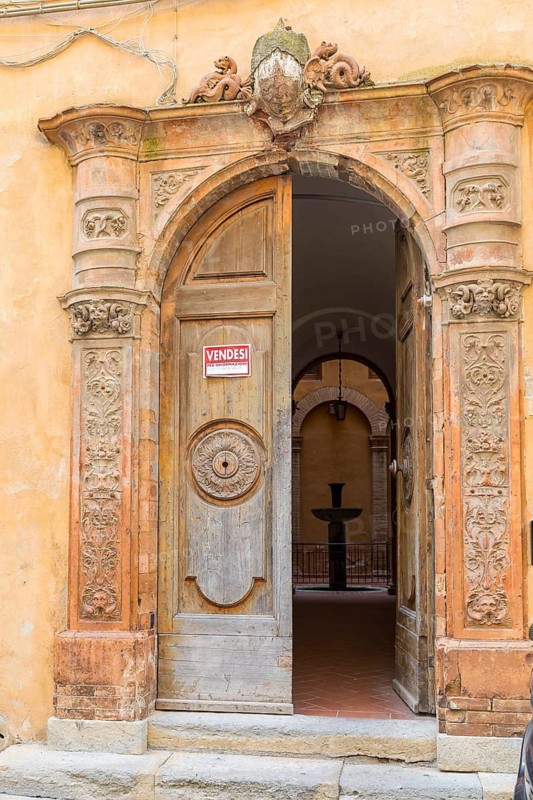 Sign VENDESI, FOR SALE on an ornate old open door with view into the courtyard with fountain, villa in the historic centre of Volterra, province of Pisa, Tuscany, Italy