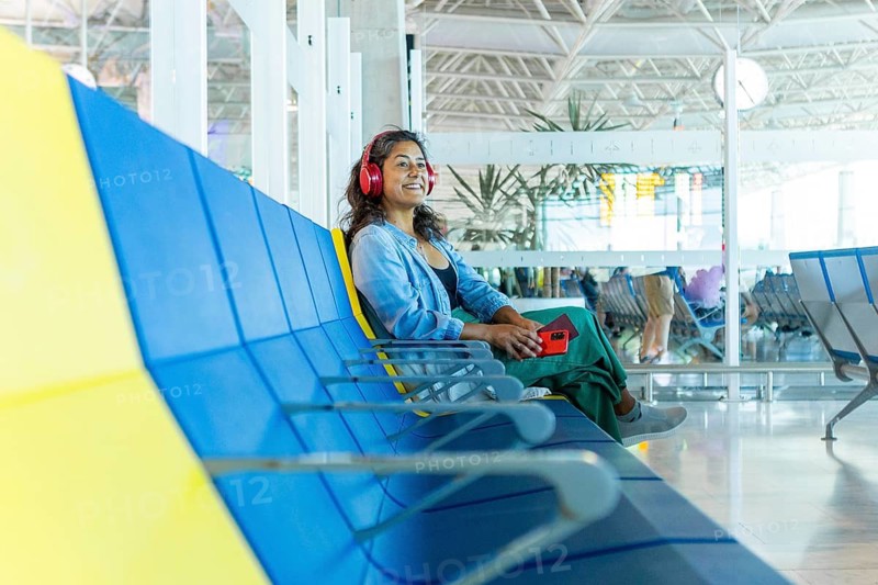A traveler enjoys a moment of relaxation in a bright airport terminal, sitting on a colorful bench Wearing headphones, she holds a phone, absorbing her surroundings