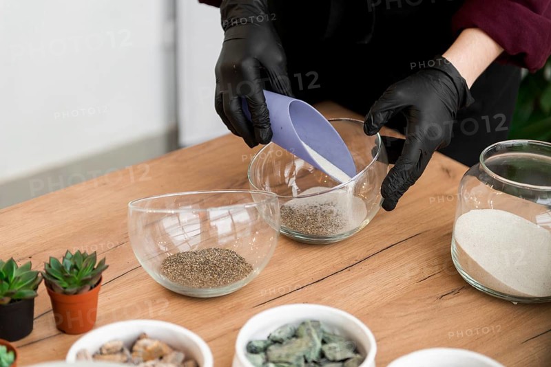 Black gloved hands skillfully pour fine sand into a glass bowl, preparing to create a terrarium. Small succulents and pebbles are ready nearby on a wooden table