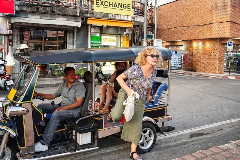 A Thai tuk tuk driver smiles as he transports tourists through the bustling streets of Chiang Rai, Thailand. A woman exits the tuk tuk, navigating the vibrant urban environment