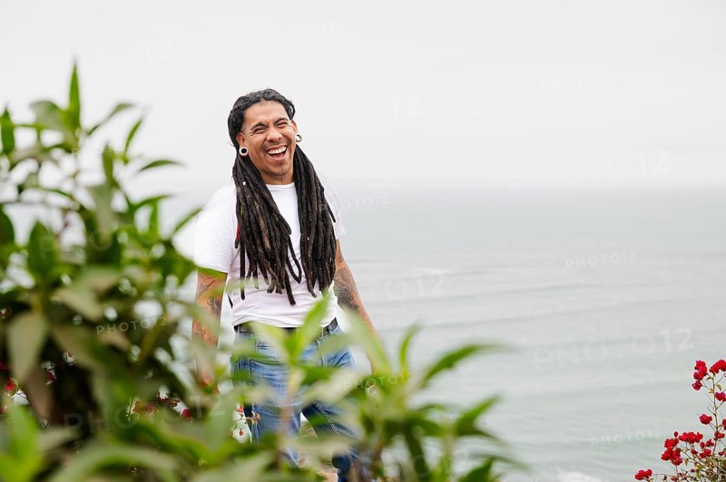 A joyful man with long dreadlocks stands near greenery with the ocean in the background, smiling broadly Captured in the vibrant Barranco district of Lima, Peru