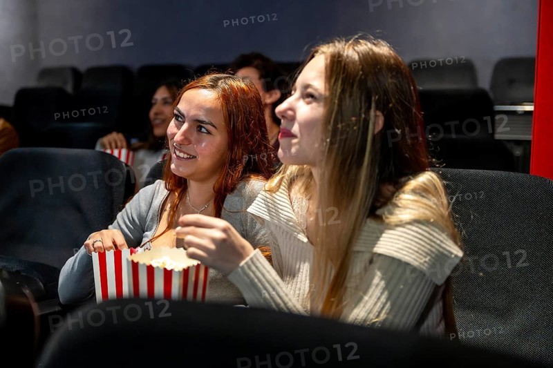 Two young women smiling and enjoying popcorn while watching a movie together in a cozy cinema, sharing laughter and a fun evening