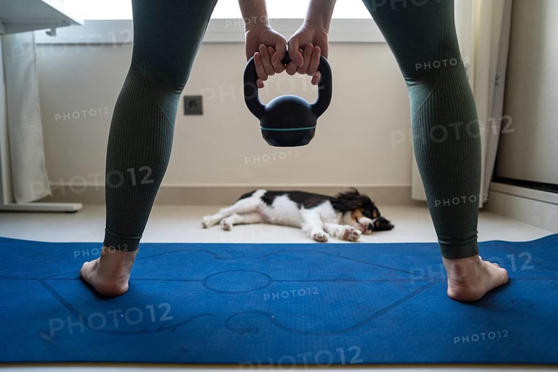 A young woman attempts to exercise at home, holding a kettlebell while her playful dog distracts her on a yoga mat. The warm, cozy setting adds charm to the scene