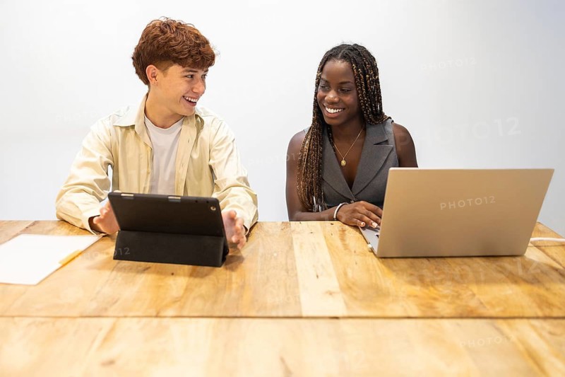 A young multiracial team collaborates enthusiastically in a coworking office. A man uses a tablet while his colleague works on a laptop, reflecting modern teamwork dynamics