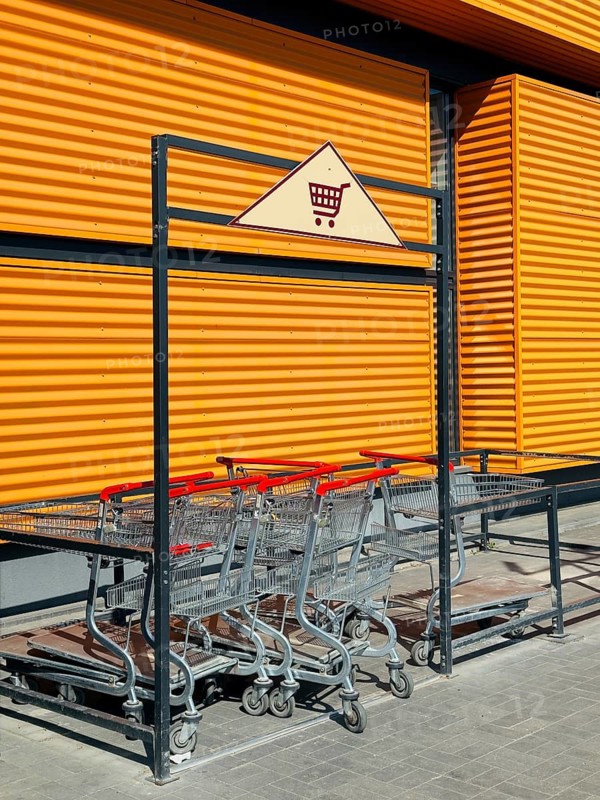 A collection of shopping carts stacked neatly in a cart corral outside a modern industrial building, featuring corrugated orange walls and a shopping warning sign