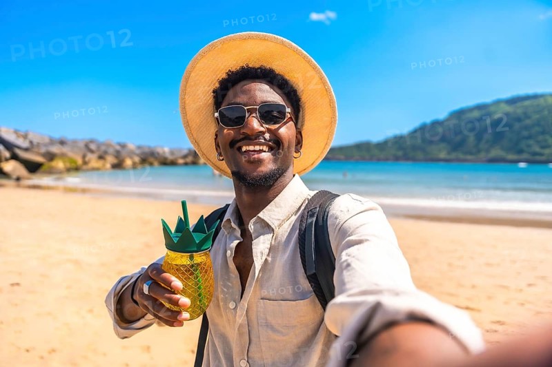 Young black man with straw hat and sunglasses enjoying a refreshing drink on a tropical beach takes a selfie