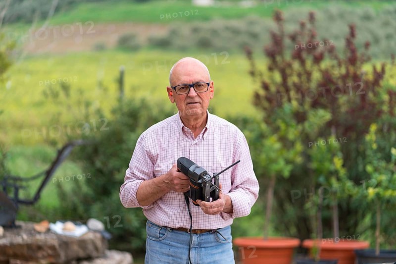 Senior man confidently holding a jigsaw with a serene natural backdrop, looking at camera, engaging in a DIY project outdoors, showcasing skill and enthusiasm in carpentry