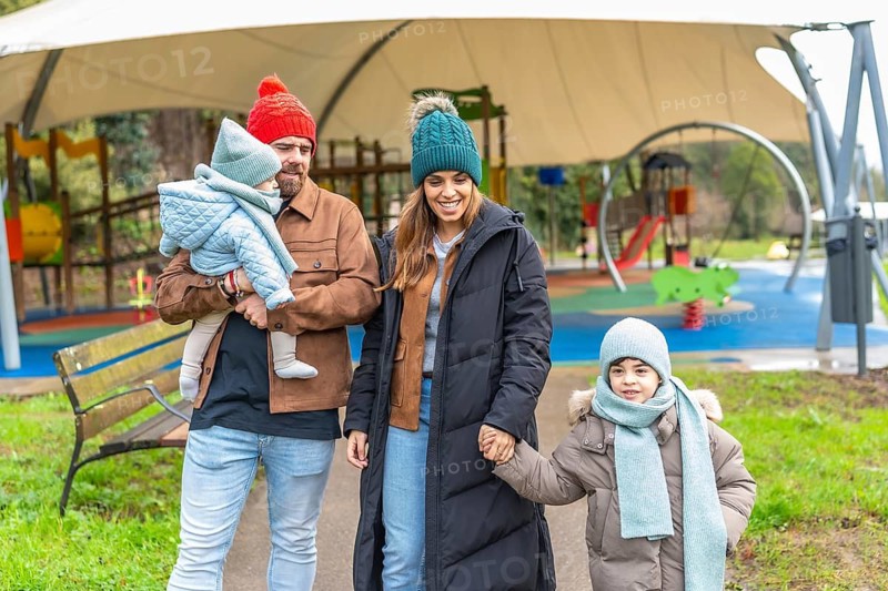 Smiling parents holding their children's hands are walking in a playground, wearing winter clothes