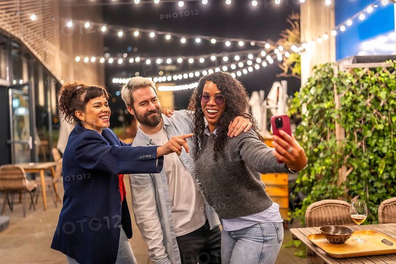 Playful multiracial friends taking selfie at night party in an outdoor terrace of a bar