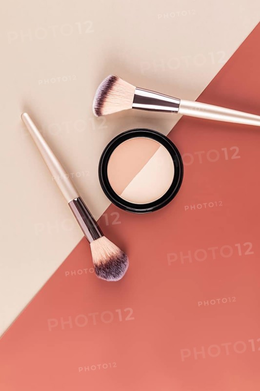 A pair of makeup brushes and a compact powder are artistically arranged on a split tone background in beige and rust, highlighting a minimalist aesthetic ideal for beauty themed projects
