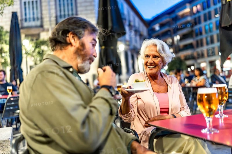 Senior man eating tapas next to partner in sidewalk cafeteria in the city of Madrid at night