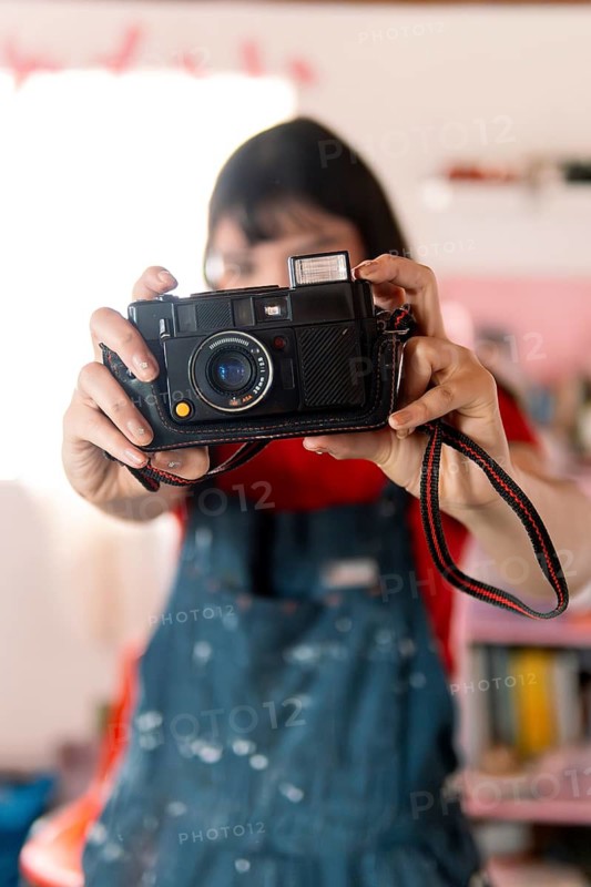 A young woman in a red shirt and paint-splattered overalls playfully holds a vintage camera, aiming it at the viewer. The creative setting and nostalgic feel highlight her artistic expression