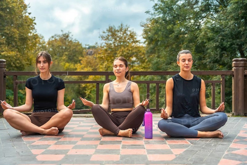 Three young women practicing yoga and meditation together outdoors in a serene park setting, finding peace, balance, and wellness while sitting in lotus position