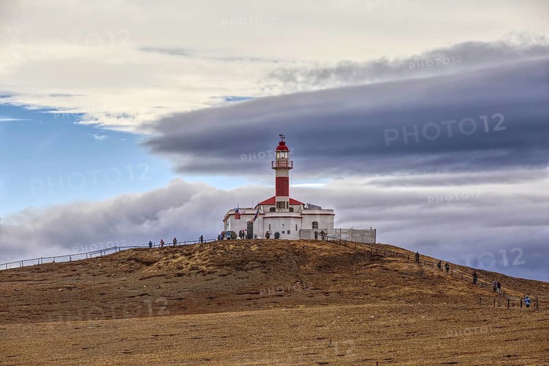 View of the lighthouse of Isla Magdelana and some tourists on a hill under a dramatically cloudy sky, Punta Arenas, Chile