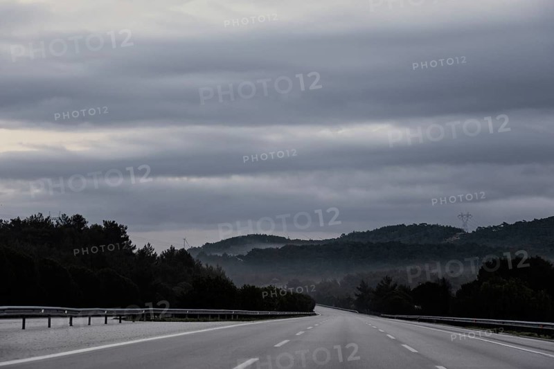 An empty highway stretches into the distance under a dramatic sky filled with thick clouds. Fog drifts around the distant hills, creating a mysterious atmosphere