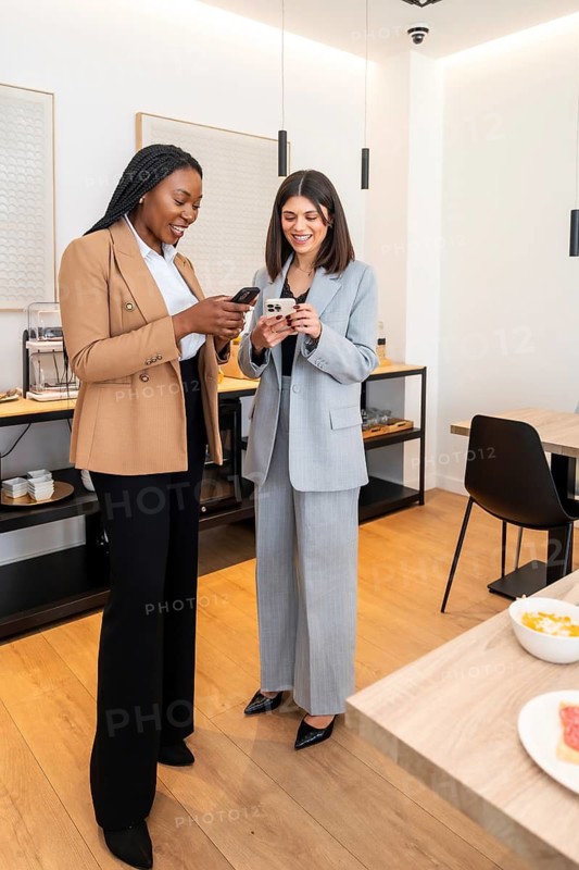 Two businesswomen are standing in a hotel lobby, using their smartphones and smiling during a business trip