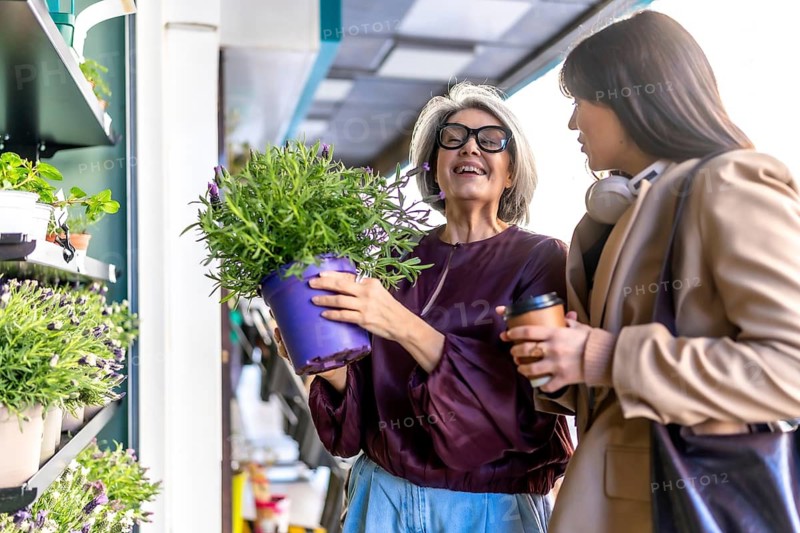 Two diverse women enjoying a joyful shopping experience at an outdoor flower shop, discussing a vibrant purple potted plant while smiling and holding a coffee cup