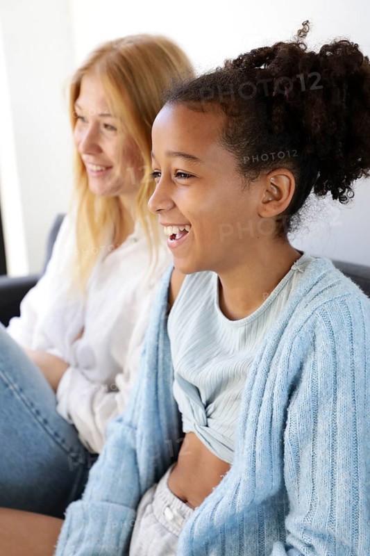 A joyful woman and girl share a laugh while sitting on a couch in a bright living room. The candid moment captures a genuine connection, showcasing happiness, togetherness