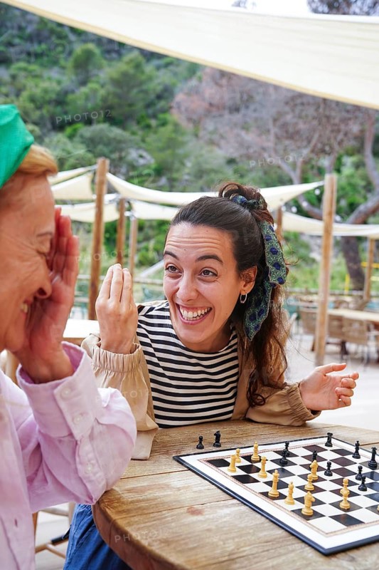 Mother and daughter share a lighthearted moment playing chess outside The scene captures smiles and strategy against a backdrop of greenery, promoting relaxation and friendship