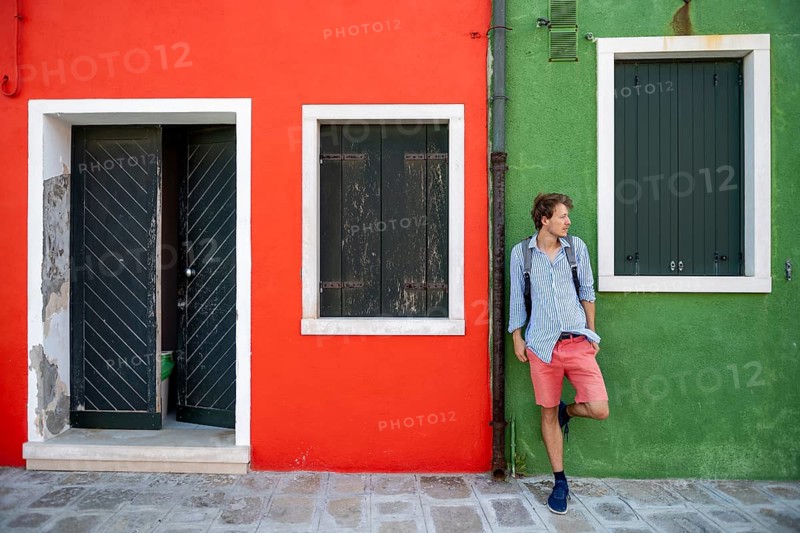 Young man with backpack leaning against a house wall, green and red house facade with entrance door and window, colourful houses on the island of Burano, Venice, Veneto, Italy