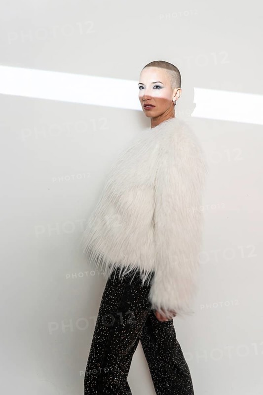 A confident middle-aged woman with a shaved head poses in a luxurious fur coat against a neutral background, showcasing modern style and elegance