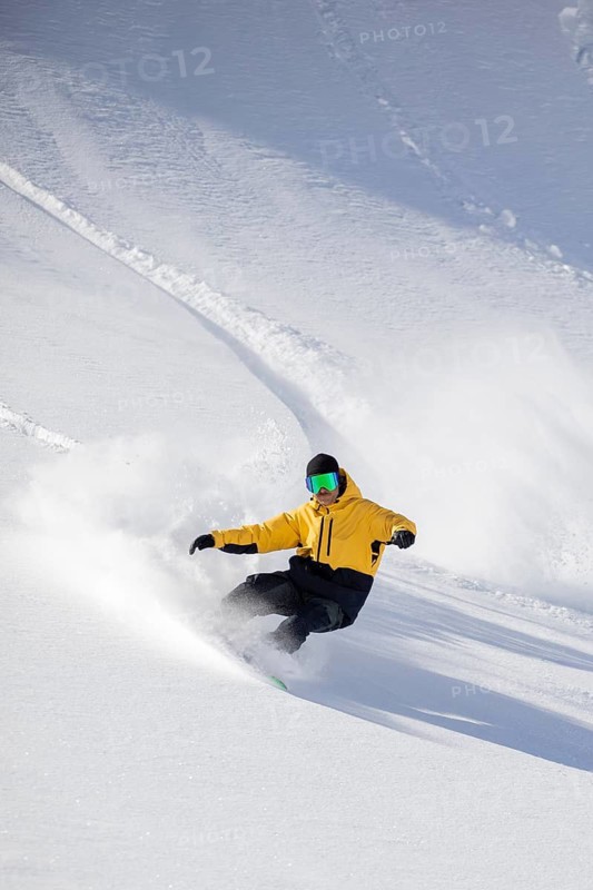 A snowboarder wearing a yellow jacket skillfully carves through fresh powder on a snowy mountain. The bright sunshine and pristine snow create a perfect winter sports scene