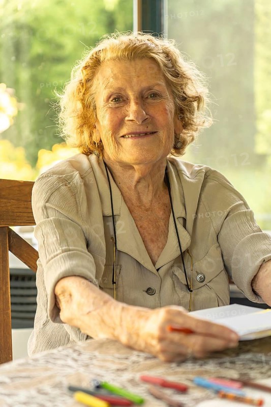 Happy elderly woman sitting at a table drawing in a notebook with colored pencils, enjoying her retirement in a bright and comfortable home environment