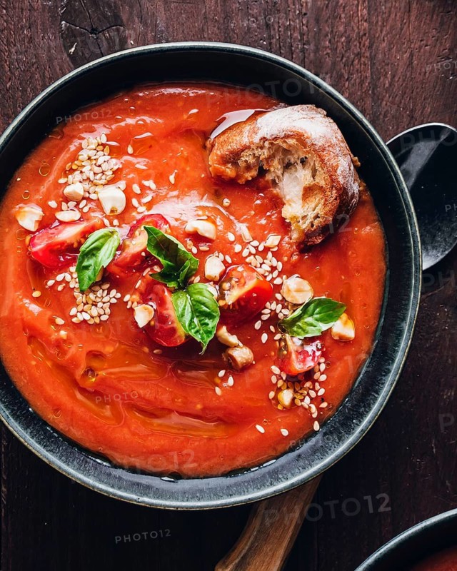 Bowl of rich tomato soup topped with cherry tomatoes, fresh basil, sesame seeds, and a piece of crusty bread on a dark wooden surface, showcasing a perfect appetizer