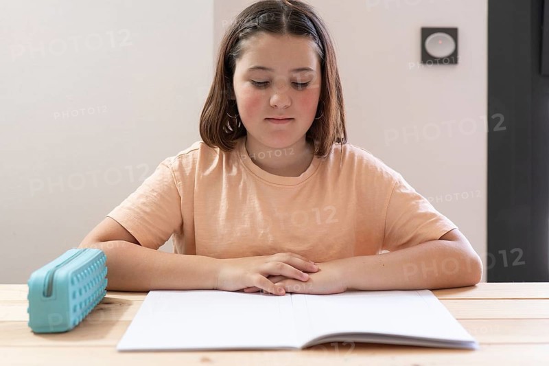 A young student in a peach shirt focuses on homework at a wooden desk with a blank notebook and blue pencil case. The setting suggests a calm, focused study environment