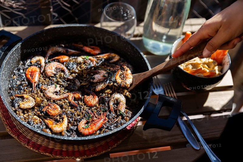A delicious black paella with shrimp and seafood, cooked with squid ink, served in a rustic pan. The dish is enjoyed outdoors with side dishes and drinks under natural sunlight