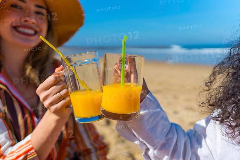 Two friends celebrating summer holidays by the sea, toasting their glasses of refreshing orange juice with straws, enjoying a relaxed moment under the sun on a sandy beach