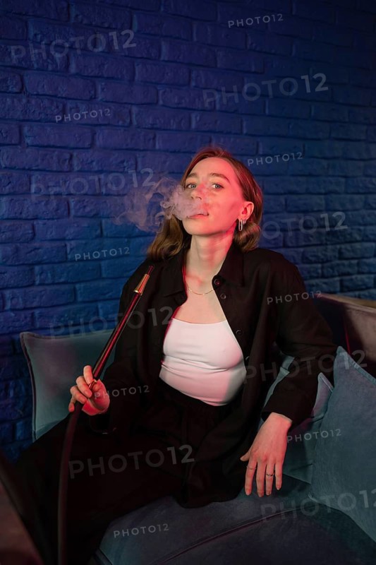 A relaxed young woman in a stylish outfit enjoys smoking hookah in a trendy lounge. She exhales smoke against a vibrant blue brick wall, creating a calm atmosphere
