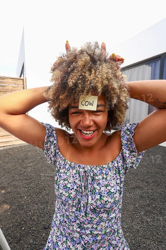 A joyful african american woman with a sticky note on her forehead labeled COW laughs while playing a fun guessing game outdoors A vibrant and cheerful moment of social interaction and play